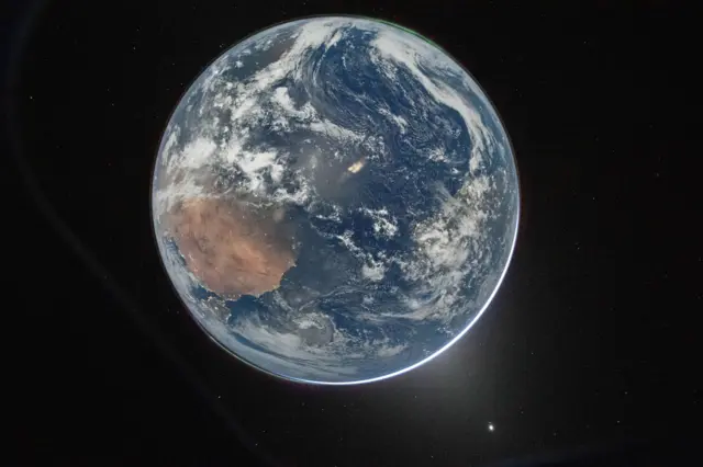 Una imagen de la Tierra desde el espacio, que sitúa el planeta en el centro contra el fondo oscuro del cosmos. Es un planeta azul y redondo. Se pueden ver nubes y una fina aurora verde en la parte superior.