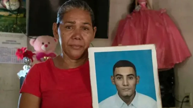 En una foto de 2024, Marcela Alvarado mira a cámara, con los ojos llorosos y una camiseta roja, mientras sostiene un retrato de su hijo detenido en el Cecot de El Salvador, José Duval Mata Alvarado.