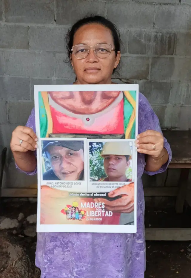 Liliana Uríes mira a cámara mientras sostiene un cartel con las fotos de su marido Israel Reyes (izquierda) y su hijo Merlon Urías (derecha).